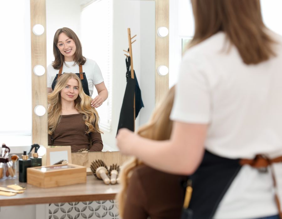 Smiling stylist and blonde client reflected in a lighted wood-framed salon mirror