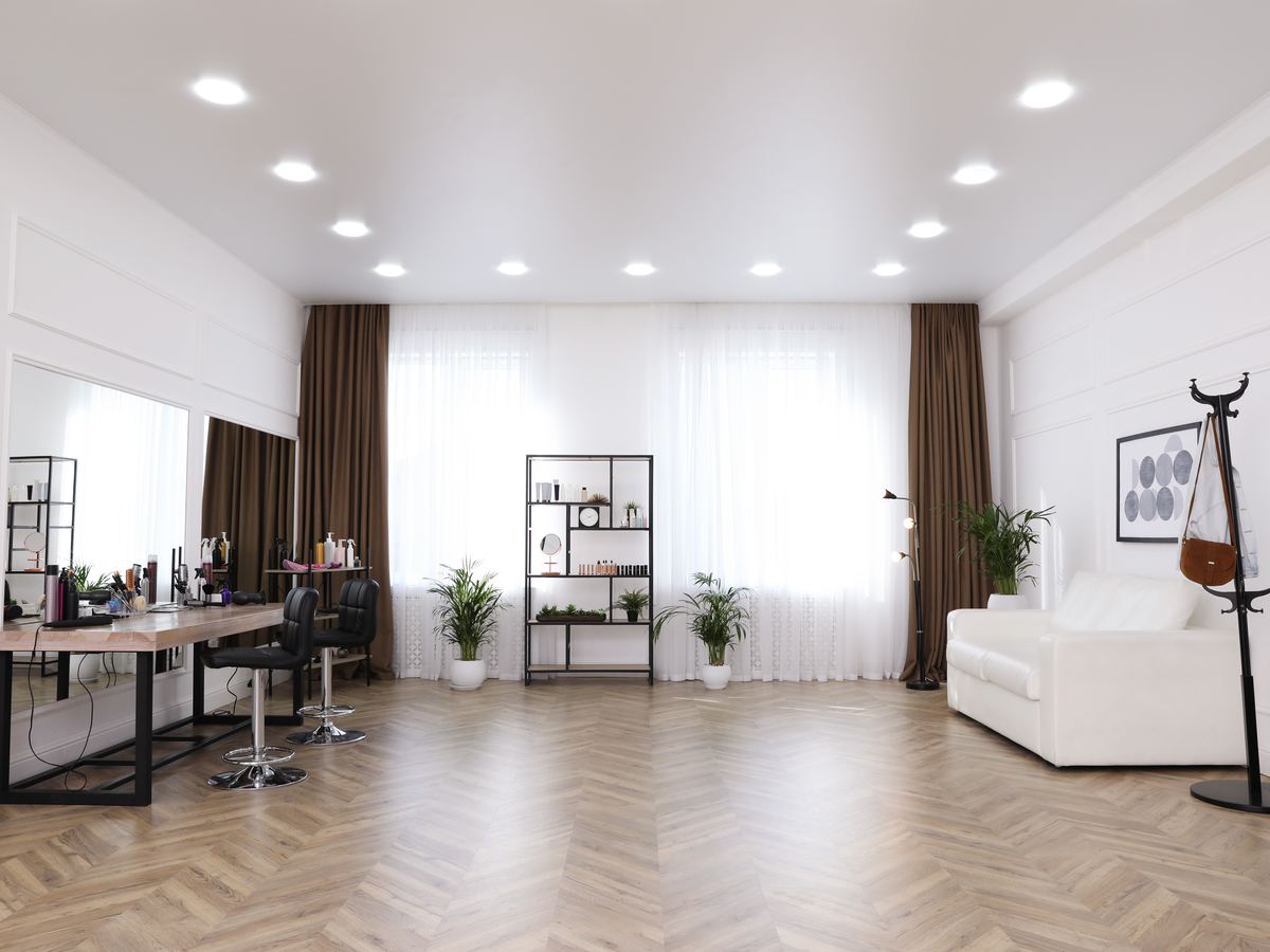 Bright modern salon interior with empty black styling chairs, light wood stations, and a white waiting sofa